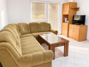 A living room with a sofa, a glass coffee table, a TV on a wooden cabinet, and a window with blinds. - Apartment Vir 1 (Vir)