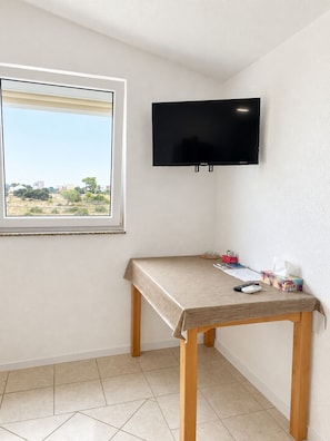 Ein Zimmer mit an der Wand befestigtem Fernseher, einem Fenster mit Blick auf Gebäude und Bäume sowie einem kleinen Tisch mit Tischdecke und einigen Gegenständen darauf.