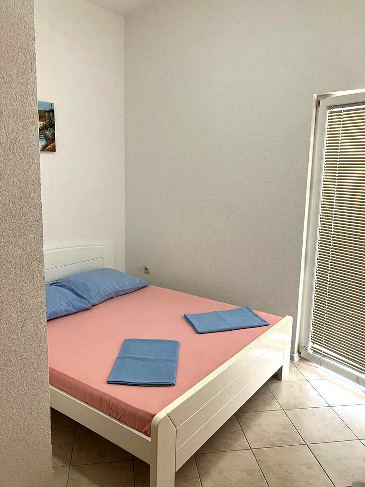 A small, single-bed room with a pink mattress, two blue pillows, and a window with blinds.