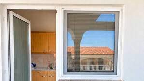Un pequeño balcón con una mesa y sillas, una ventana con persianas, y vistas a un edificio con tejado rojo.