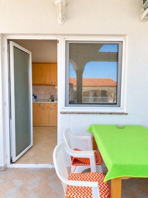 Ein kleiner Balkon mit Tisch und Stühlen, ein Fenster mit Jalousien und Blick auf ein Gebäude mit rotem Dach.