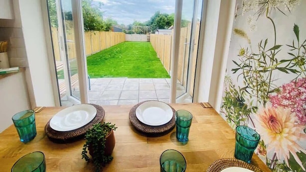 A dining area with a table set for two, a view of a green lawn through a glass door, and a floral wall decoration. - Twickenham House Sleeps 7 with Parking - Isleworth (Greater London)