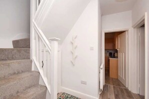 Una escalera con peldaños alfombrados conduce a un rellano con una pared blanca y un adorno. Una puerta da a una zona de cocina con armarios de madera y una encimera.
