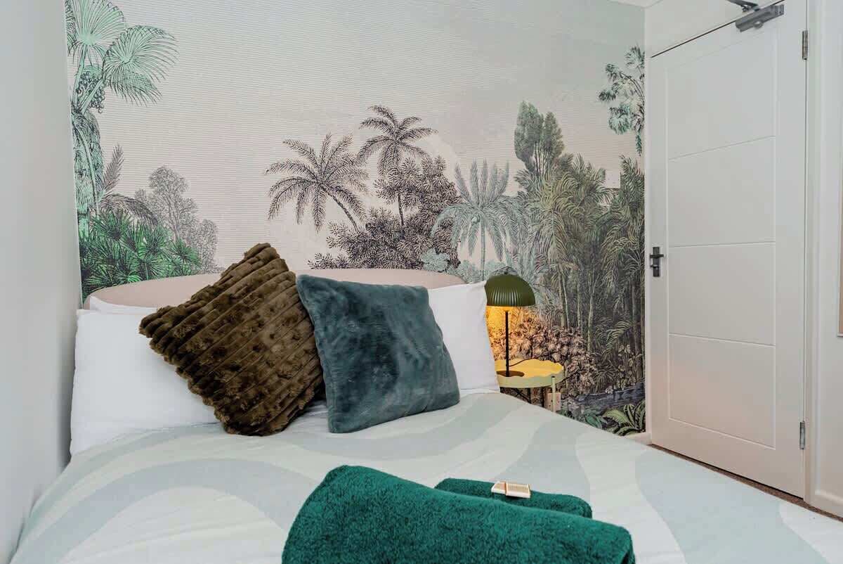 Une chambre à coucher avec un lit, des oreillers, une couverture verte et une fresque murale représentant un paysage tropical.