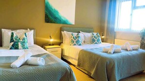 Una habitació d'hotel amb dos llits, cada un amb un coixí i una tovallola enrotllada. Hi ha una tauleta de nit amb una làmpada, un quadre a la paret i una finestra amb cortines.