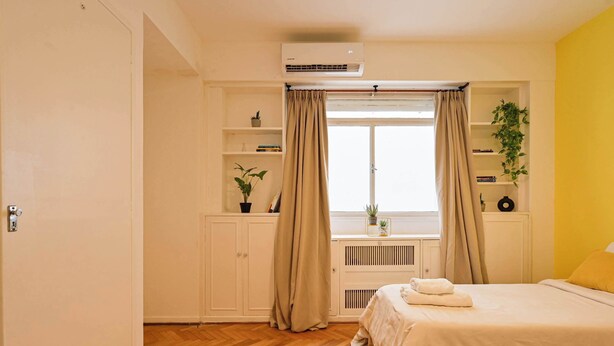 A bedroom with a bed, a window with curtains, a radiator, and a shelf with plants. - MOT  Rentals + Laprida 1300 - 3 Bd 2 Ba in Recoleta! (Buenos Aires)