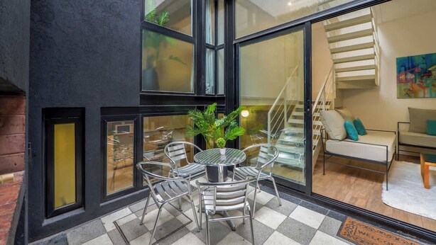 Un interno moderno con una porta in vetro che dà su una scala, una zona pranzo con sedie in metallo e un tavolo rotondo, e un soggiorno con divano e cuscini.