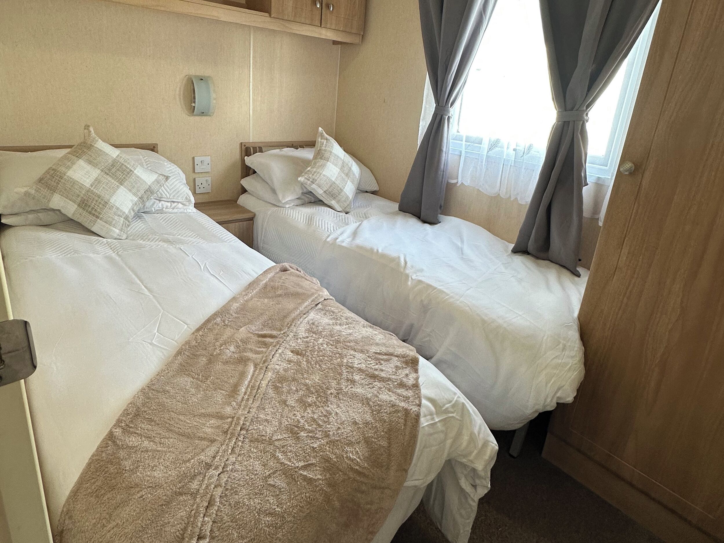 Two beds with white linens and plaid pillows, a wooden headboard, a nightstand, a wall-mounted light, and a window with curtains.