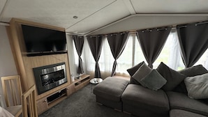 A modern living room with a flat-screen TV, built-in fireplace, grey sofa, and large windows with curtains. - C43 The Hideout (Abergele)