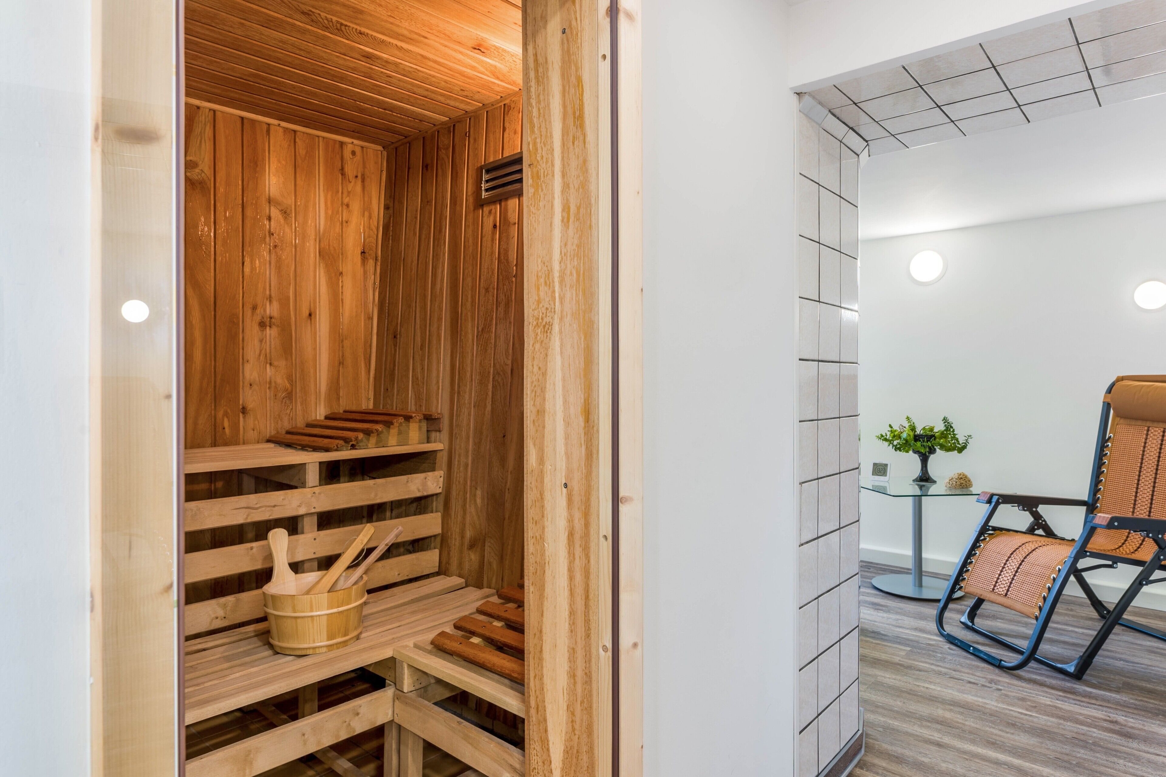 Un sauna avec des bancs en bois et un seau en bois, un mur carrelé et une chaise avec un siège en osier tressé.