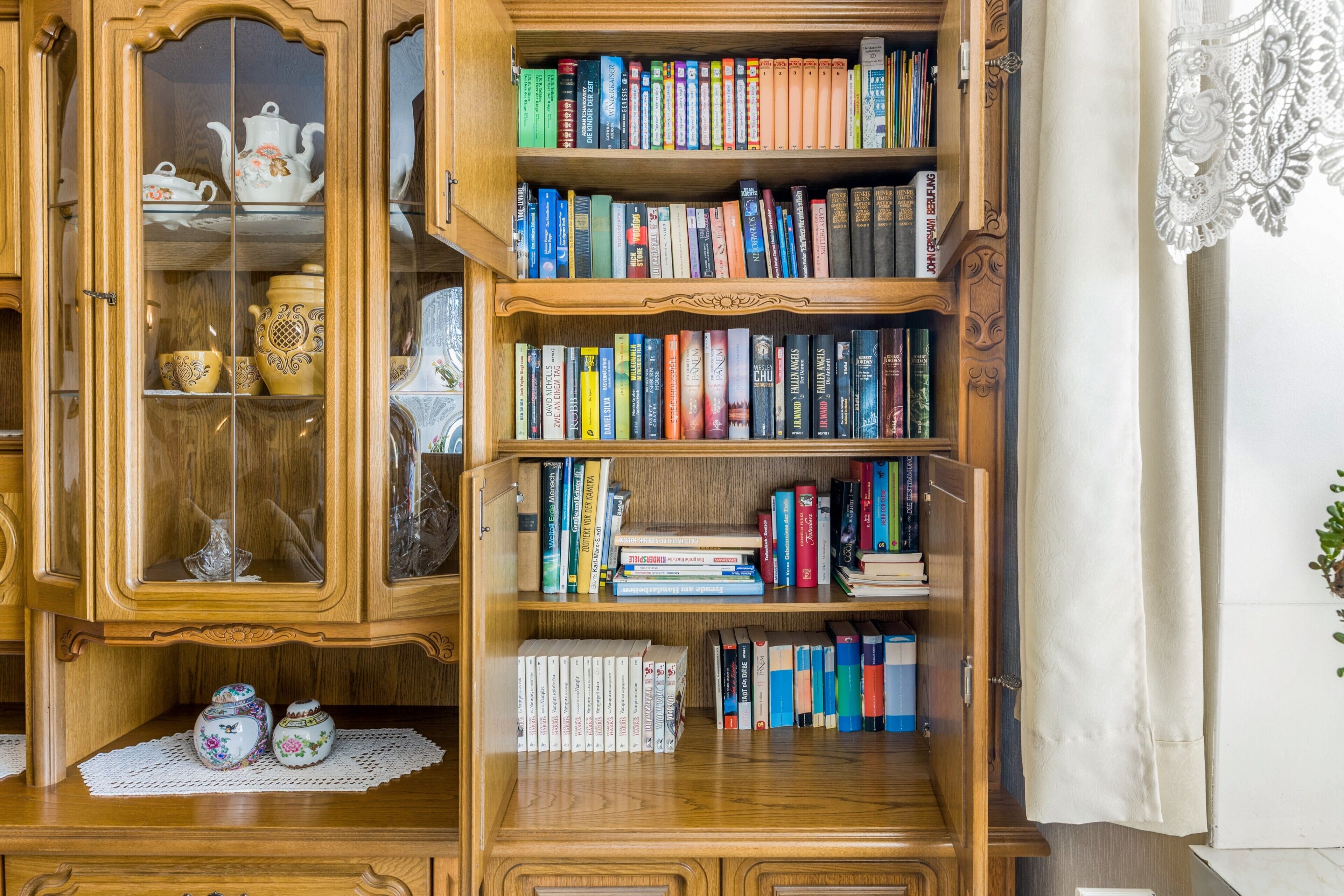Ein Bücherregal aus Holz mit mehreren Regalböden, gefüllt mit Büchern, ein Kabinett mit Glastür und dekorativen Gegenständen sowie ein Spitzenvorhang.
