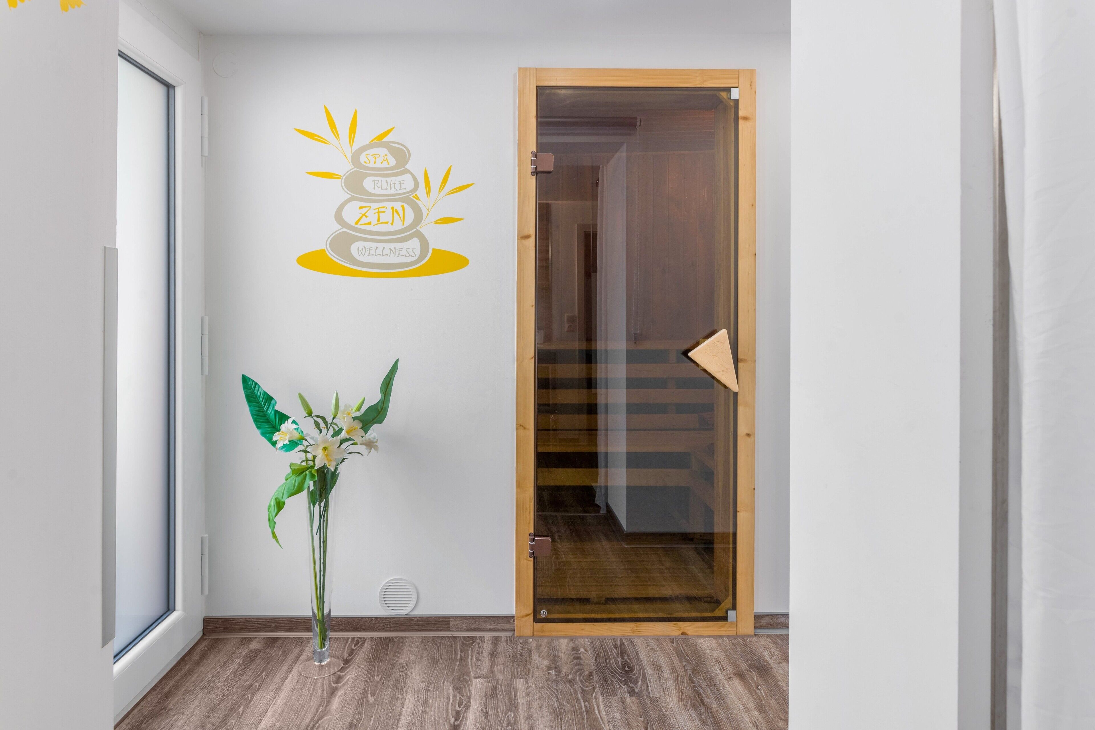 Un couloir avec une porte en bois, une enseigne fixée au mur portant les mots « SPA » et « ZEN WELLNESS », et une plante en pot avec des fleurs blanches et des feuilles vertes.