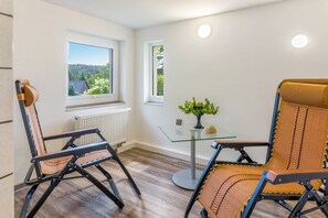 Une pièce avec deux fauteuils à bascule, une petite table sur laquelle est posé un vase de plantes, et une vue sur une forêt par la fenêtre.