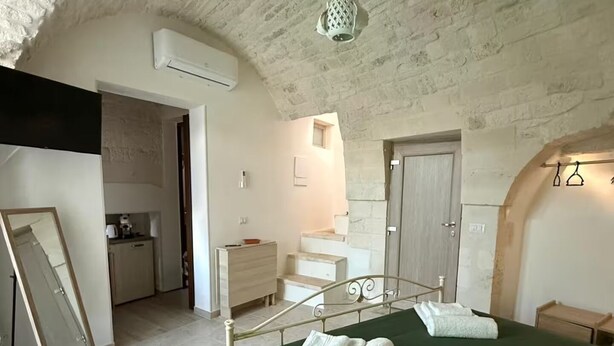 Une chambre avec une couvre-lit verte, une serviette blanche et un lit à cadre métallique. Un petit escalier mène à une salle de bain, et un appareil de climatisation blanc est fixé au mur.