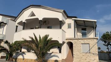 A modern two-story house with a balcony, stone base, and palm trees in the foreground.