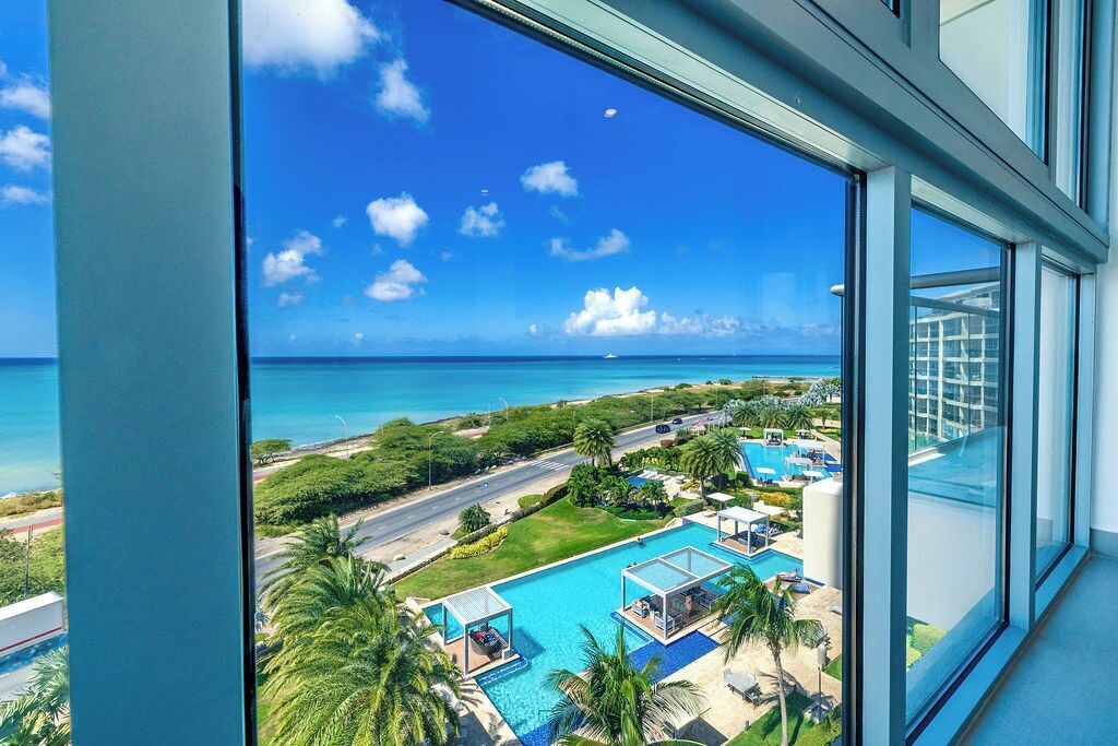 Blick von einem Hotelzimmer mit großem Fenster auf einen Swimmingpool, üppiges Grün und einen strahlend blauen Himmel.
