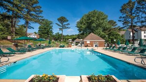 Una piscina rectangular con indicador de profundidad de 6 pies, rodeada de sillas de descanso verdes y sombrillas, con un pequeño edificio de ladrillo y casas al fondo.