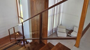 A modern staircase with wooden steps and a glass railing, leading to a living area with a sofa and curtains. - Luxury house in contact with nature and private pool in Praia do Forte (Açu da Torre)