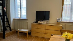 Una habitación acogedora con suelo de madera, un televisor sobre un mueble de madera, una mesa de comedor con sillas y una ventana con cortinas translúcidas.