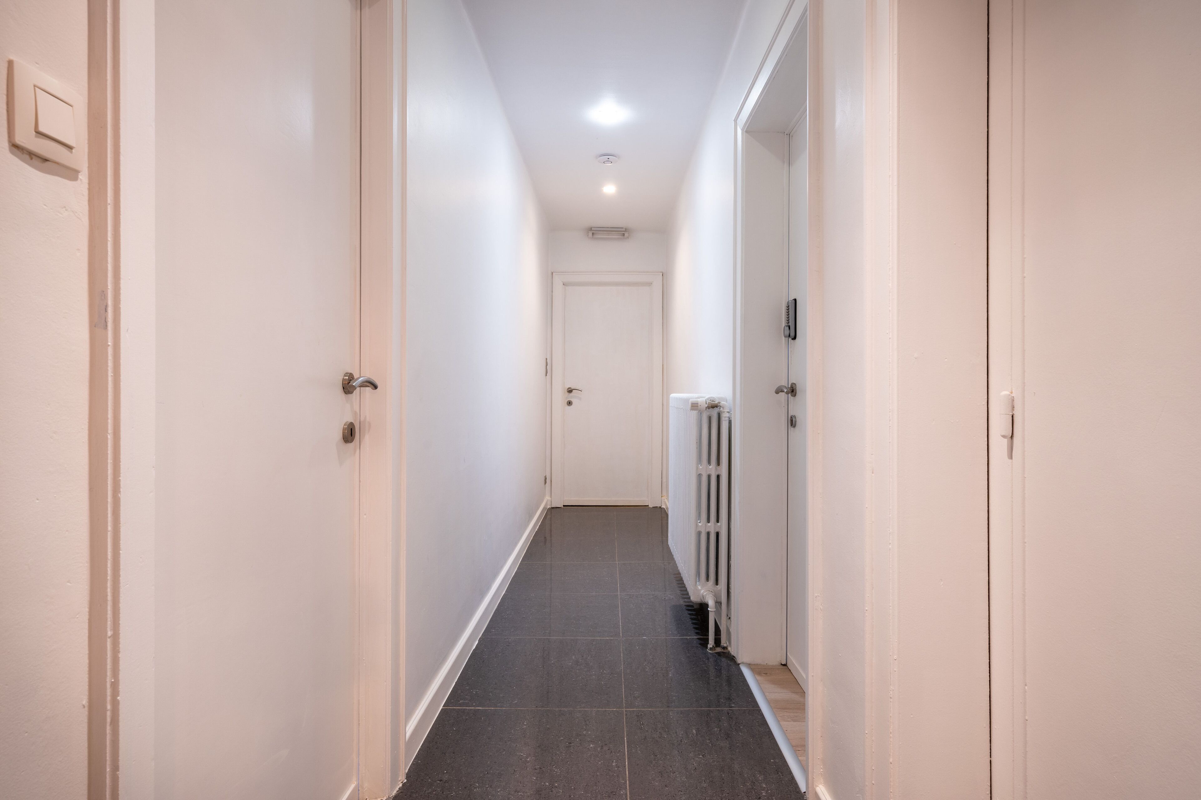 A narrow hallway with white walls, a dark tiled floor, and a white door at the end.