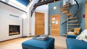 A modern living room with a fireplace, a blue sofa, a wicker chair, and a spiral staircase. - The Rose Suites (Bruges)