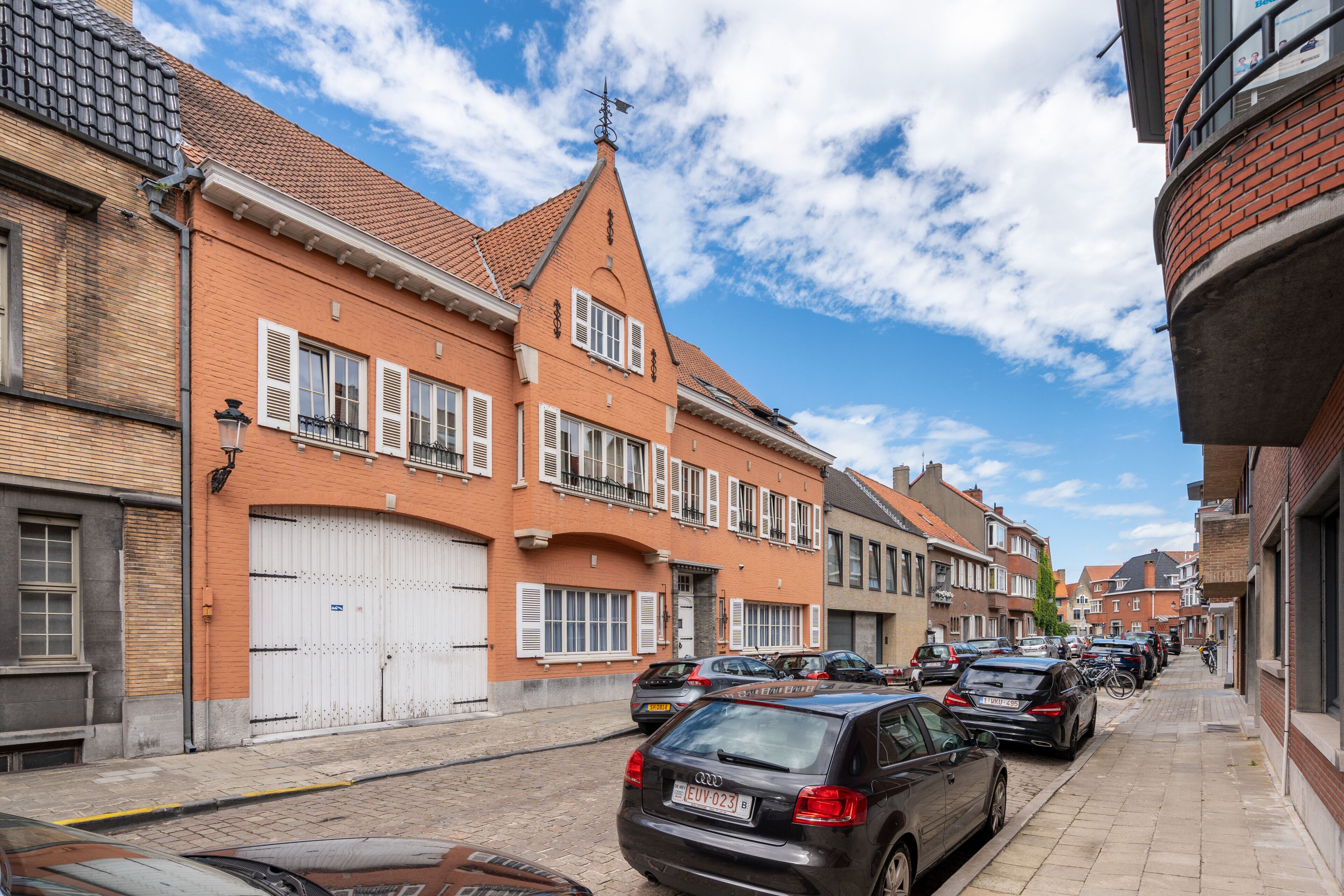 A narrow street lined with brick buildings, parked cars, and a clear blue sky.