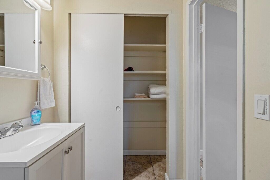 Un baño con lavamanos blanco, espejo y toallero. Se ve una puerta que da a un armario con repisas.