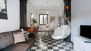 Un salon avec un sol à carreaux, un canapé, un fauteuil transparent, un téléviseur et un coin repas avec une table et des chaises.