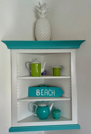 Une étagère à la couleur turquoise, ornée d’un ananas, avec deux théières vertes, un pichet violet et un panneau turquoise sur lequel on peut lire « PLAGE ».