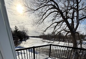 Une vue depuis un balcon doté d’une balustrade, donnant sur une rivière, avec de la neige au sol et des arbres en arrière-plan.