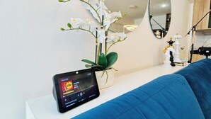 Ett modernt vardagsrum med en blå soffa, ett vitt konsolbord, en smartphone som visar en musikapp, en orkidé i en kruka och en stor oval spegel.