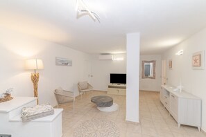 Een moderne woonkamer met een flatscreentelevisie, een rond hockertje, een witte bank en een eethoek met een witte tafel en stoelen.