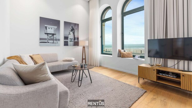 A modern living room with a grey sofa, a coffee table, a TV on a wooden stand, and a large window with a view of the sea. - Beach Loft - luxury apartment with sea view in Ostend (Oostende)