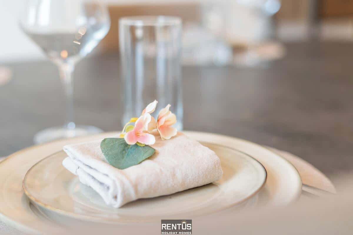 Ein ordentlich gefaltetes Handtuch mit einer Blume auf einem Teller, ein Glas Wasser und ein hohes Glas in einer Essecke.