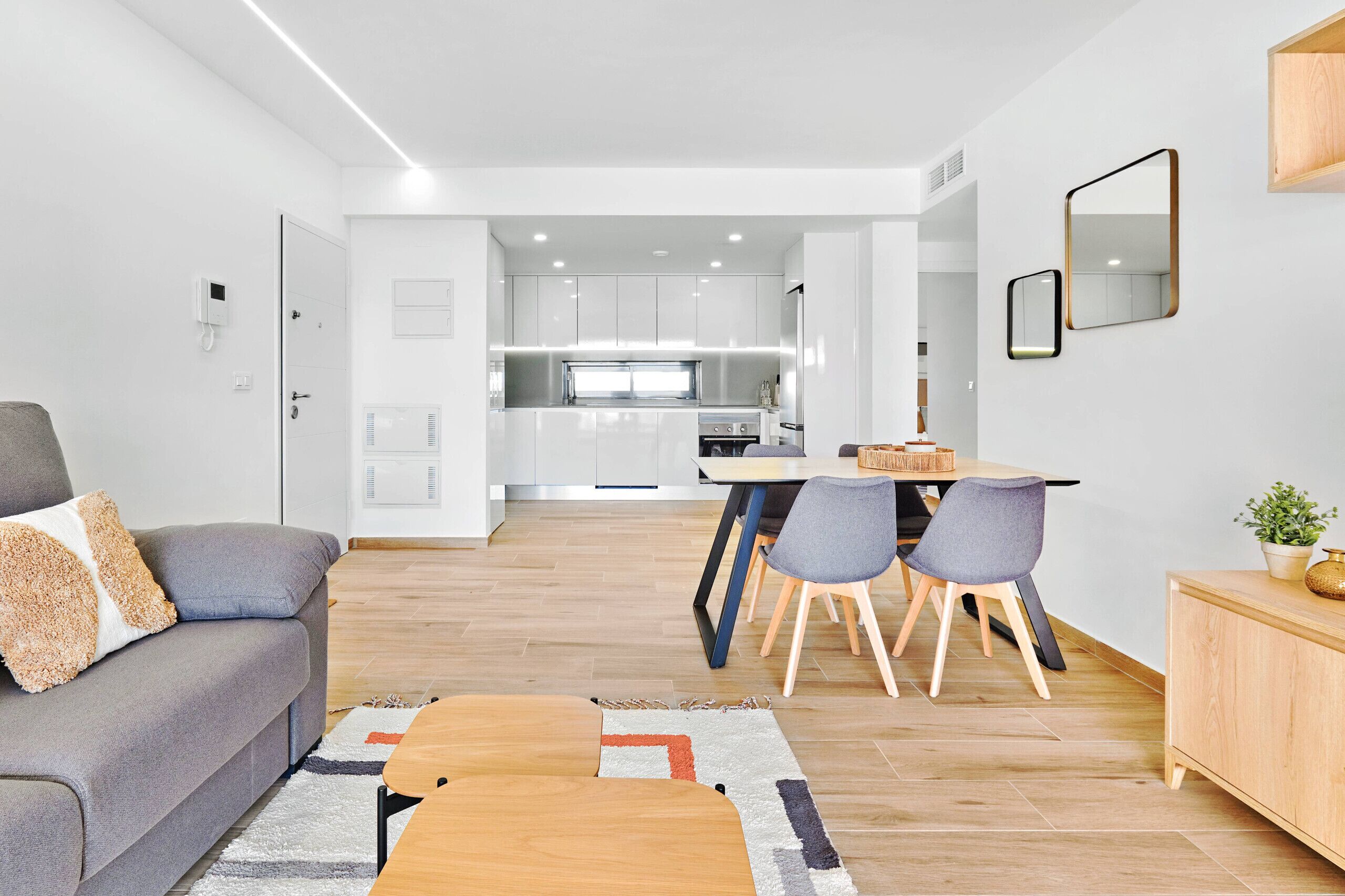 A modern living room with a grey sofa, a wooden coffee table, and a dining area with a table and chairs. A kitchen is visible in the background with white cabinets and a microwave.