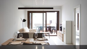 Un'area pranzo moderna con un tavolo apparecchiato per due persone, una divano sullo sfondo e una porta scorrevole in vetro che dà su una zona salotto esterna.