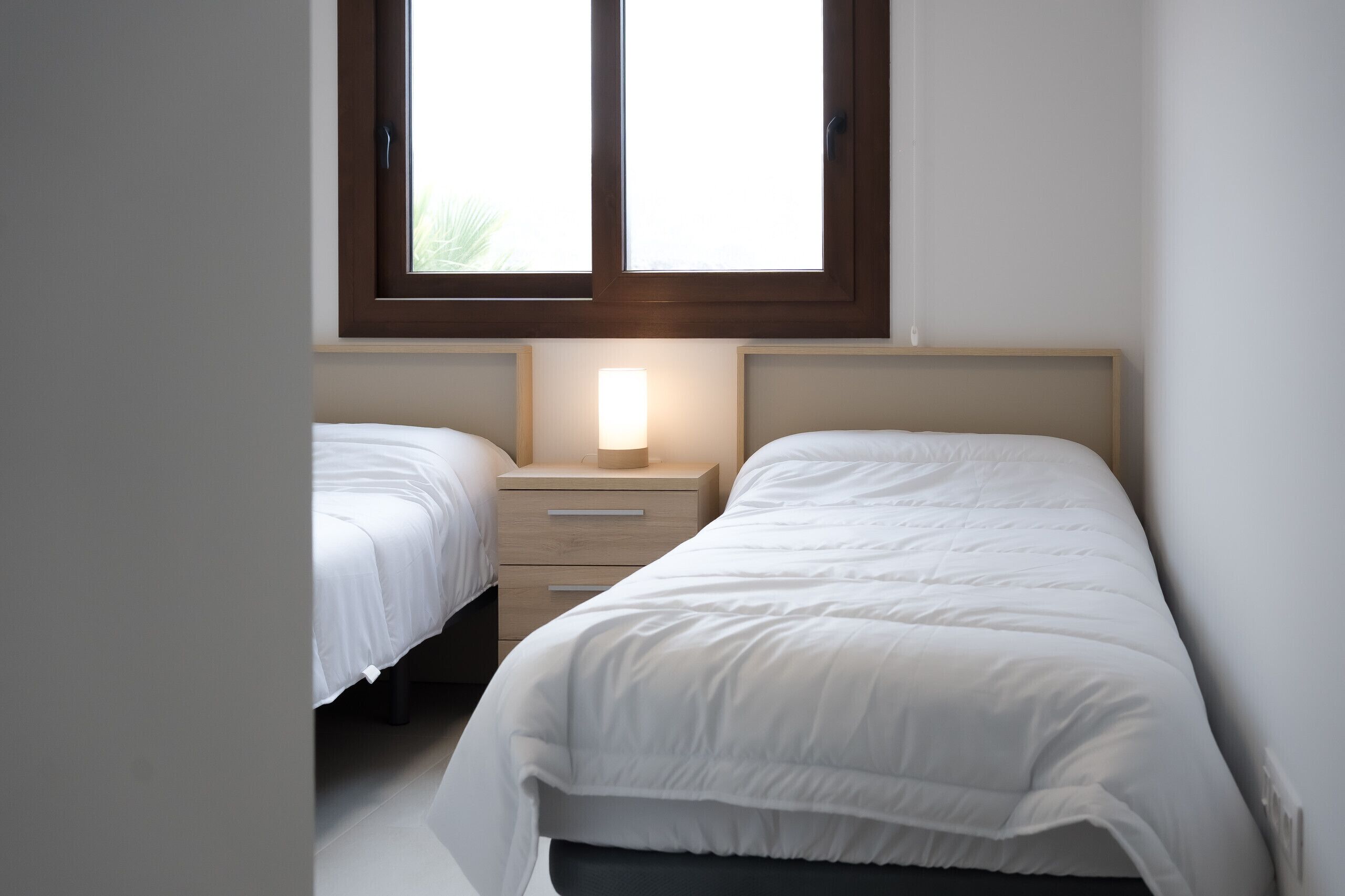 A bedroom with two single beds, a bedside table with a lamp, and a window above the beds.