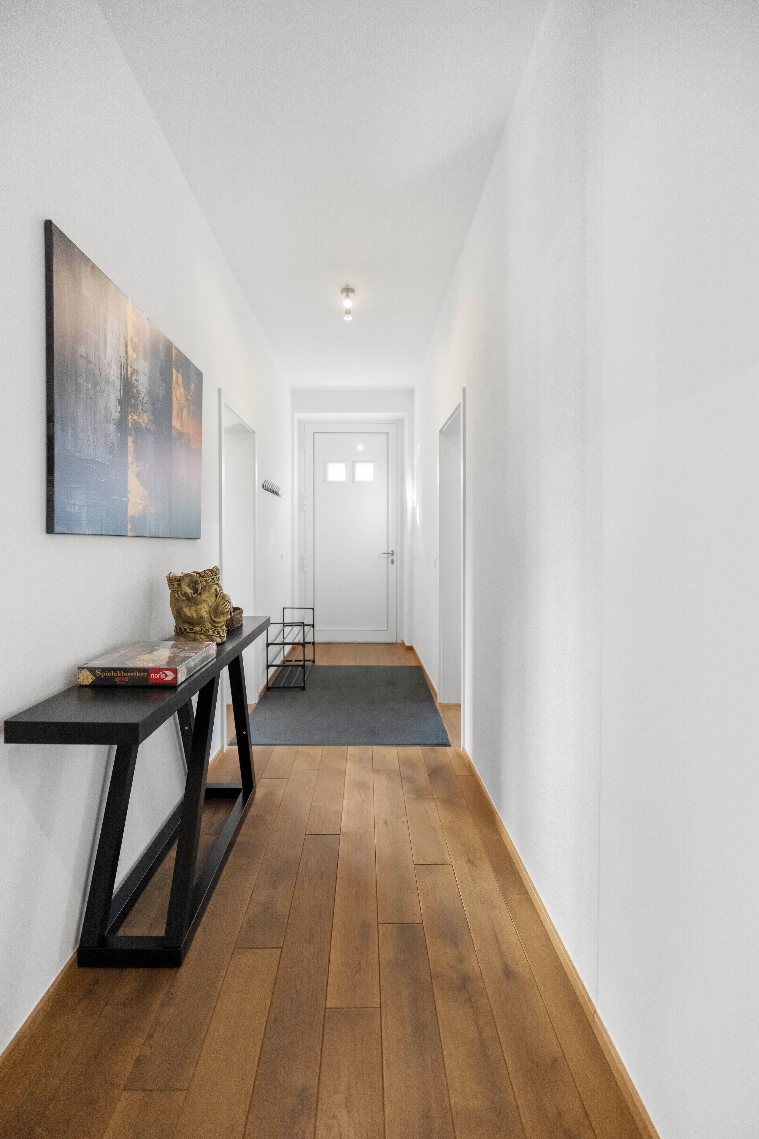 Un couloir doté d’un plancher en bois, une console noire ornée d’un objet décoratif, un tableau encadré et une porte au fond.
