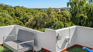 Un balcon avec une chaise en métal, une petite table et une vue sur un paysage verdoyant, avec des arbres et une étendue d’eau au loin.