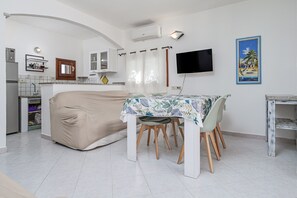 Ein kompakter Wohnraum mit Küchenzeile, einer Essecke mit Tisch und Stühlen, einem Schlafplatz, einem an der Wand befestigten Fernseher und einem Strandbild.