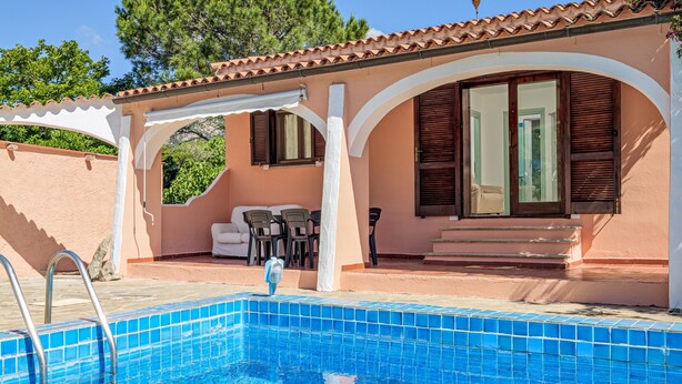 Une piscine avec un bord carrelé, un patio couvert doté de meubles d’extérieur, et un bâtiment avec un toit en tuiles et des volets en bois.