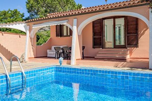 Une piscine avec un bord carrelé, un patio couvert doté de meubles d’extérieur, et un bâtiment avec un toit en tuiles et des volets en bois.