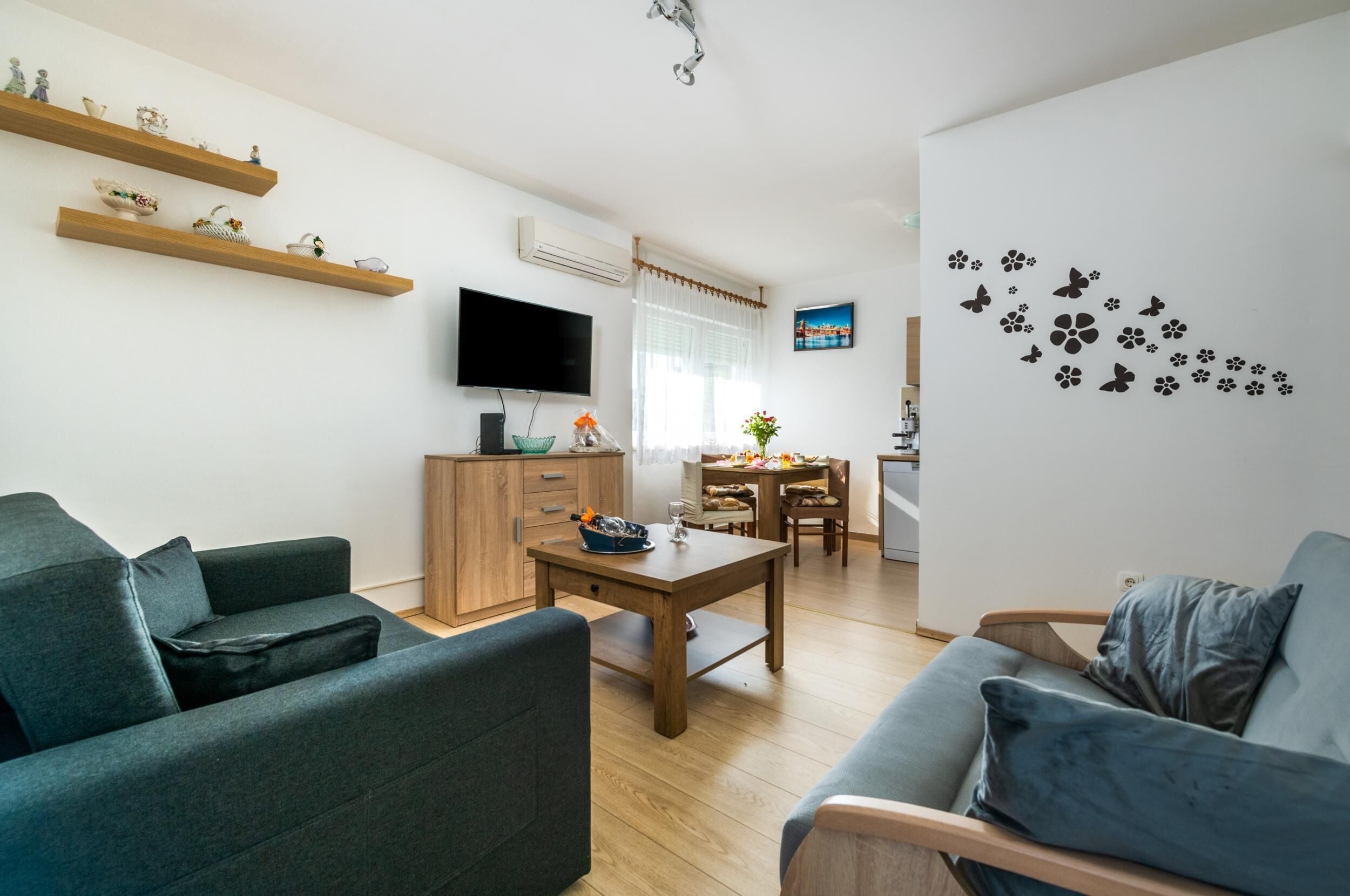 Ein modernes Wohnzimmer mit einer Couch, einem Couchtisch und einem an der Wand montierten Fernseher. Darüber befinden sich Holzregale über der Couch und im Hintergrund ein Esstisch mit Stühlen.