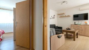 Una sala de estar moderna con un sofá, una mesa de centro y un televisor. Se observa una puerta de madera y una ventana con cortinas.