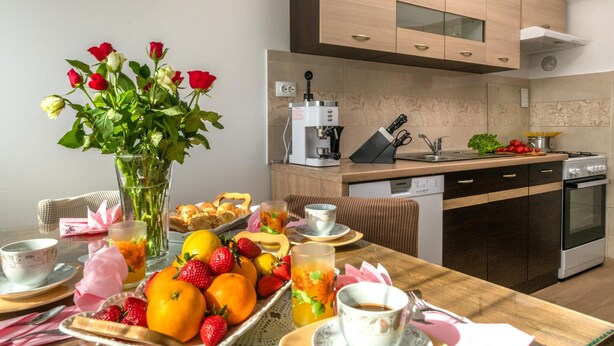 Una mesa de desayuno con frutas, pasteles y flores, una cafetera y una cocina con electrodomésticos modernos.