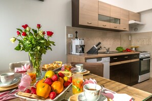 Ein Esstisch mit Früchten, Gebäck und Blumen, eine Kaffeemaschine sowie eine Küche mit modernen Geräten.