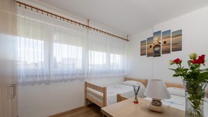 Un dormitorio con una cama de madera, una mesita de noche con lámpara, una ventana con cortinas traslúcidas y una pared decorada con cuadros.