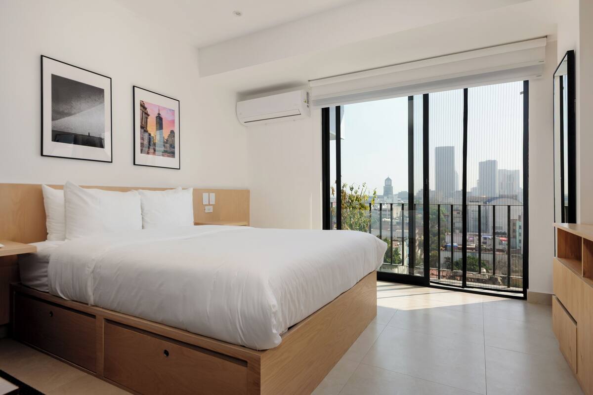 Ein modernes Schlafzimmer mit einem großen Bett, einem Holzkopfende, einem Nachttisch und Blick auf die Stadt durch einen Balkon mit schwarzer Geländer.