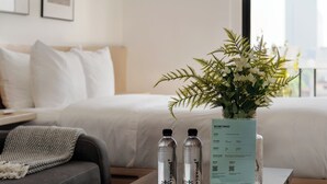 A hotel room with a bed, a grey couch, a wooden coffee table, a vase with white flowers, two water bottles, and a welcome card. - Via Insurgentes by Wynwood House (Mexico City)
