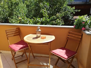 Ein runder Holztisch mit einer Laterne und einem Glasobjekt, zwei Holzsessel mit roten Kissen sowie ein Balkon mit Fliesenboden und Grünpflanzen im Hintergrund.